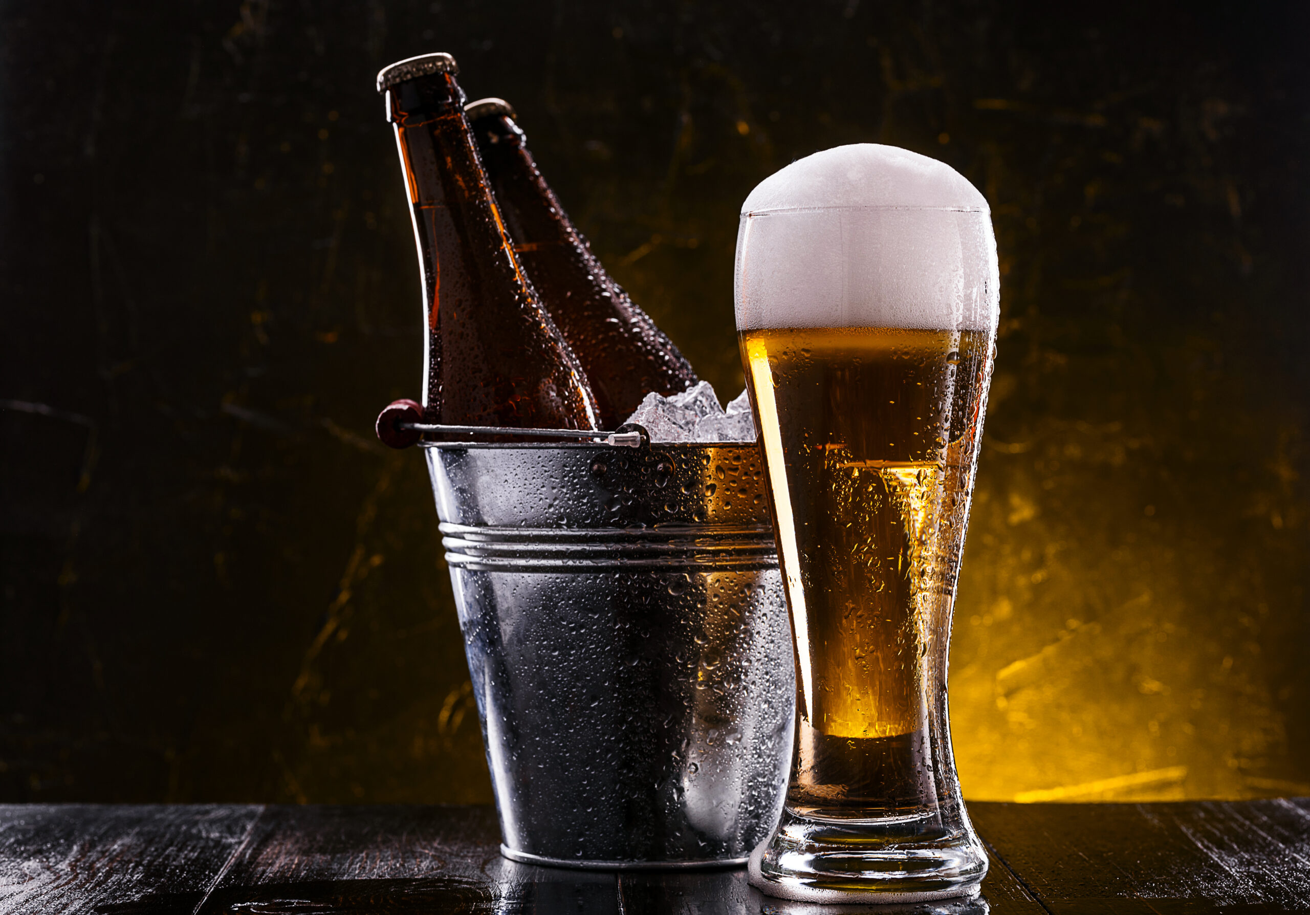 two bottles of beer in a bucket with ice and a glass of beer with lush foam next to a dark background